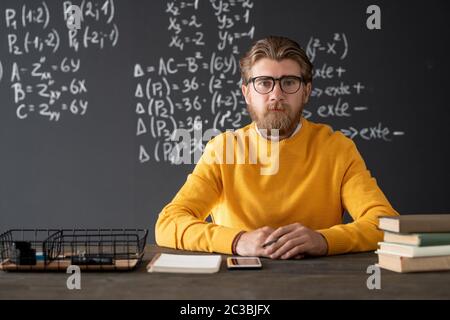 Young bearded teacher of algebra sitting by table against blackboard with formulas and equations during online lesson in classroom Stock Photo