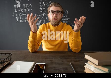 Confident teacher of algebra explaining new subject to his online students while sitting by table with books and notepad in front of camera Stock Photo
