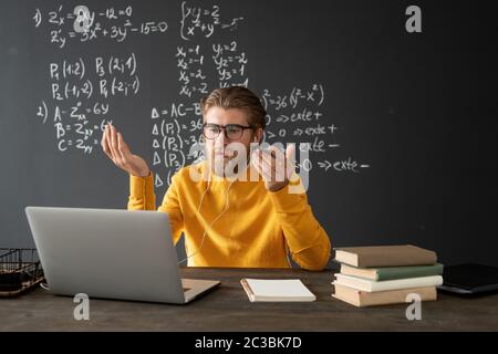 Confident teacher in eyeglasses looking at his audience on laptop display while explaining new formula on blackboard during online lesson Stock Photo