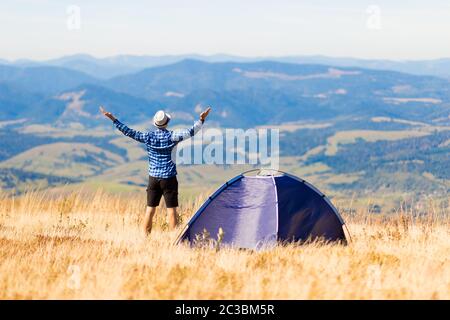Back view of happy traveler man on top of mountain raised hands enjoying freedom and view near tent camping outdoor. Travel adventure lifestyle succes Stock Photo