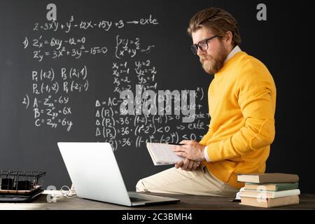 Young contemporary teacher with copybook sitting on table in front of laptop and talking to his online audience about algebraic formulas Stock Photo