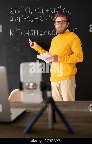 Bearded chemistry teacher in casualwear pointing at chemical formula on blackboard while looking at smartphone camera during online lesson Stock Photo