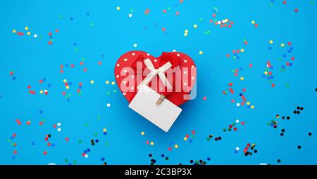 closed red heart-shaped cardboard box and empty white business card on a clothespin on a blue background with multi-colored shiny confetti, festive ba Stock Photo