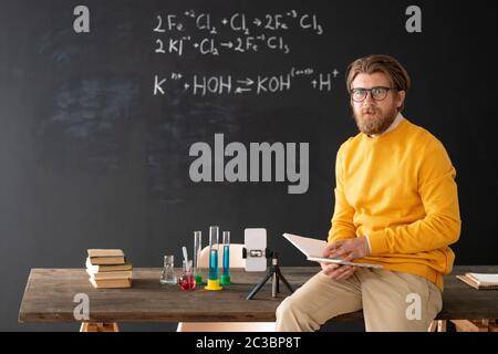 Confident bearded chemistry teacher with open book sitting on table against blackboard and preparing lecture for online students Stock Photo