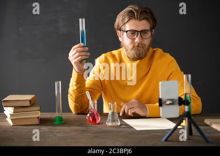 Confident chemistry teacher in casualwear showing flask with blue fluid to his online audience while explaining its characteristics at lesson Stock Photo