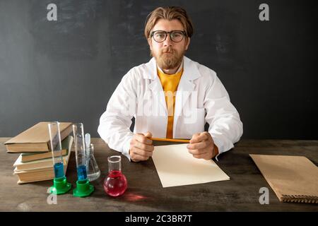 Young confident teacher of chemistry in whitecoat looking at his online audience while sitting by table against empty blackboard Stock Photo