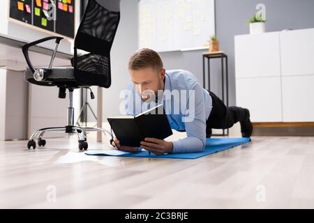 Businessman Reading Notes While Doing Plank Exercise In Office Stock Photo