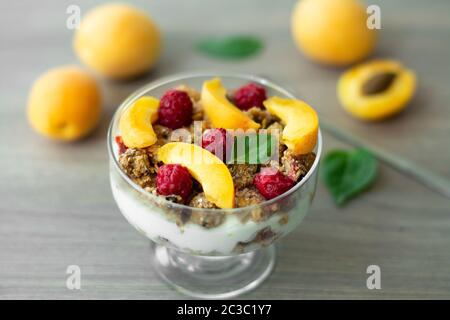 Healthy parfait with greek yoghurt, homemade granola, slices of apricot and raspberries in a glass goblet. Grey wooden background with apricots Stock Photo