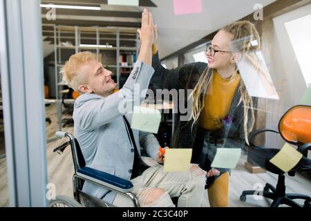 Business concept, successful disabled people at work. Happy successful young man in wheelchair giving a high five to his pretty female coworker with Stock Photo