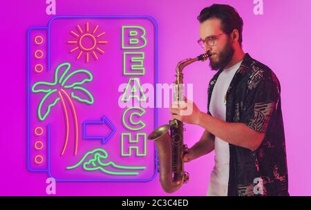 Young jazz musician playing the saxophone on gradient studio background in neon light with sign BEACH. Concept of music, hobby, festival. Joyful inspired artist, flyer. Summertime, sale, vacation. Stock Photo