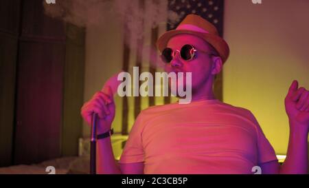 Hipster man smoking hookah and dansing in bright color neon illumination. Funny smoker holding shisha pipe has fun alone in dark room Stock Photo