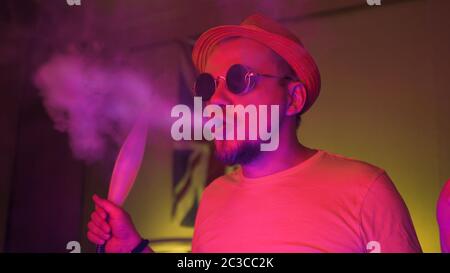 Positive bearded man smokes hookah in pink neon illumination in dark home room. Copy spase for text at both sides Stock Photo