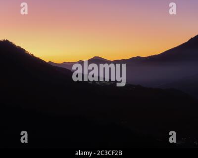 Highlands and houses glowing in the distance against the backdrop of a mountain range and sunset sky Stock Photo