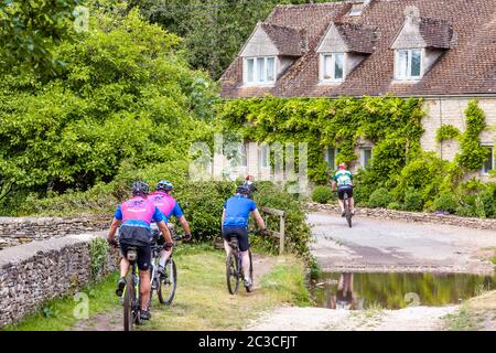 Cyclists passing cottages beside the ford in the Cotswold village of Duntisbourne Rouse, Gloucestershire UK Stock Photo