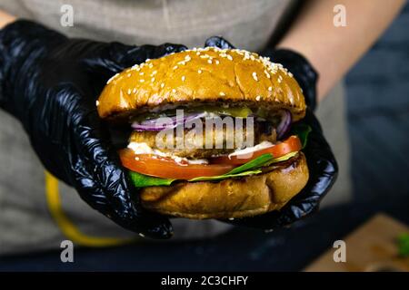 Woman's hands in black rubber gloves are holding juicy bun burger with meat cutlet, lettuce, tomato, shredded cheese and marinated cucumber. Top views Stock Photo