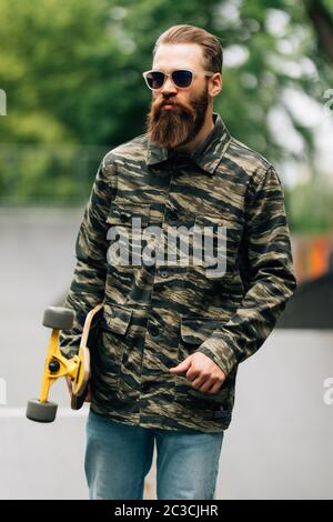 Bearded man holding longboard or skateboard in the park . Stock Photo