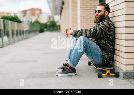 Young bearded man sitting on his skateboard in a skate park. Stock Photo