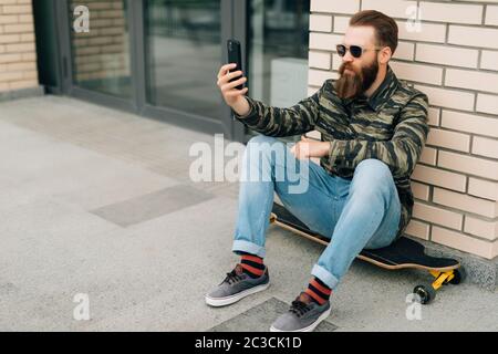 Young hipster man taking photo with his phone sitting on the longboard Stock Photo