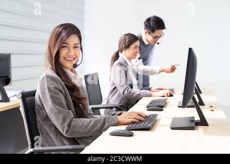 Young adult friendly and confidence operator woman agent smiling with headsets working in a call center with her colleague team working as customer se Stock Photo