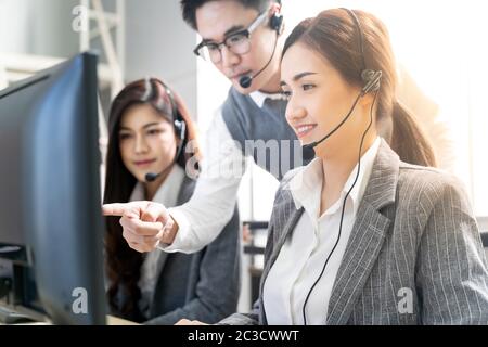 Young adult confidence with smiling supervisor teaching to his team of operator team agent with headsets and desktop computer in a call center to moni Stock Photo