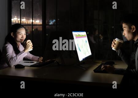 Young adult asian businessman and woman drinking hot cofee while working late at night in their office with desktop computer and laptop. Using as hard Stock Photo