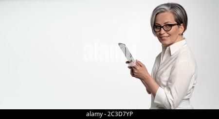 Cheerful business woman works on digital tablet on white background. Mature lady in white looking at camera. Horizontal blank with empty spase for Stock Photo