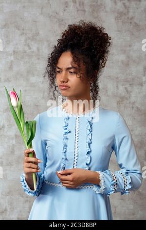 Dissatisfied Young African girl in a blue dress with a Tulip on a gray background. The concept of women's day and spring Stock Photo
