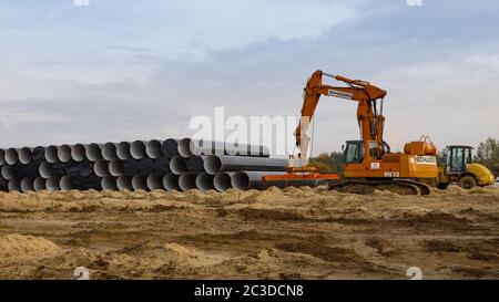 Stack of 8-m-sections of gas pipeline tubes next to a working site. A yellow caterpillar with a pipe handling clutch is standing by. Stock Photo