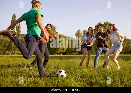 Young friends playing football game on green meadow. Students having fun on summer weekend in nature Stock Photo