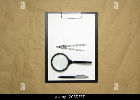 A tablet with a white sheet of A4 format with magnifier, pen and divider on a beige craft paper. Concept of analysis, study, attentive work. Stock photo with empty place for your text and design. Stock Photo