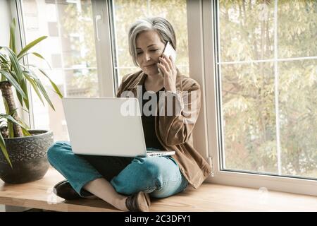 Charming woman talking on cell phone while working on laptop. Gray-haired aged woman sits cross-legged on wide windowsill, leaning her back against Stock Photo