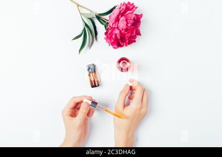 Female hands apply cosmetic serum next to oil and flower on white background, overhead view. Stock Photo