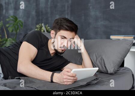 Lazy young bearded man lying on bed and using tablet while wasting time during coronavirus quarantine Stock Photo