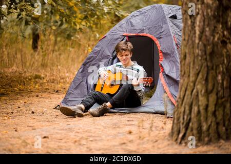 Smiling young man sitting near touristic tent and playing guitar in autumn forest Stock Photo