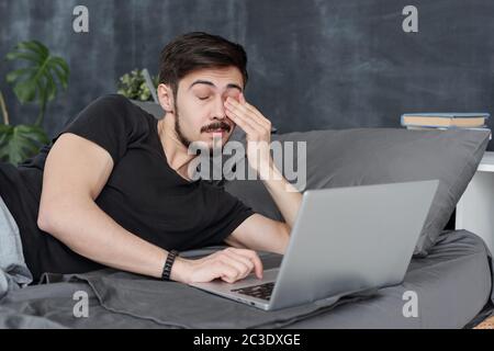 Tired young man having eye fatigue lying on bed with laptop and rubbing eye Stock Photo