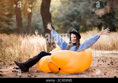 Young woman in blue sweater and black hat admires forest sunset outdoor with hands up on inflatable sofa Stock Photo