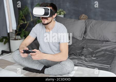 Smiling young man sitting with crossed legs on bed and playing video game using joystick and virtual reality headset Stock Photo
