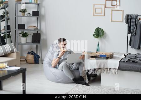 Young man sitting in bean bag and eating pizza while watching series online in quarantine Stock Photo