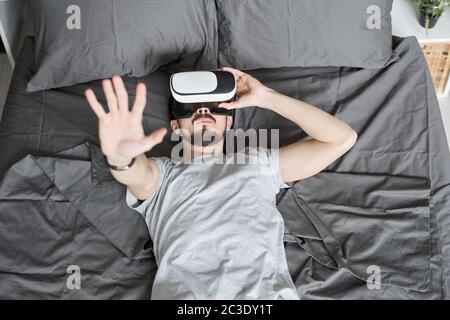 Above view of young bearded man immersed into virtual reality lying on bed and watching video in VR goggles Stock Photo