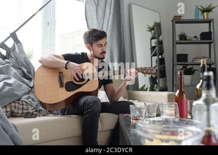 Young bearded man sitting on sofa with curtains and playing guitar in flat after party Stock Photo