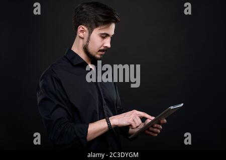 Serious young bearded man standing against black background and analyzing data on tablet Stock Photo