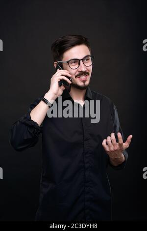 Cheerful confident young manager in eyeglasses gesturing hand while talking to client by phone against black background Stock Photo