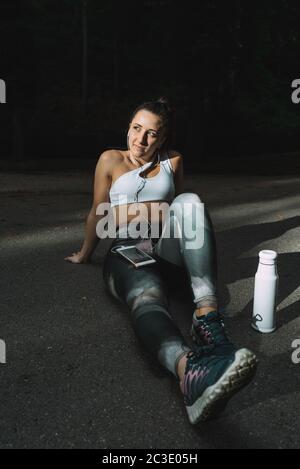 Pretty runner girl resting on the floor while listening to music on her smartphone with a bottle of water. Exhausted concept, free time and sports. Stock Photo