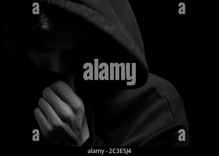 a young guy in a black hoodie on a black background with his face covered. Stock Photo