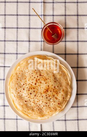 Top view of stack of fresh pancakes on plate with small bowl of flower honey standing near by on checkered tablecloth Stock Photo