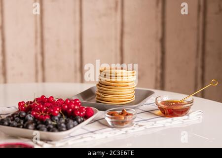 Stack of small homemade pancakes on plate, fresh ripe red currant and blackberries, small glass bowls with honey and almond kernels Stock Photo