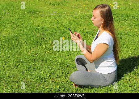 A young beautiful girl of European appearance in a white T-shirt with long blond hair sits on green grass, on the lawn with a mobile phone in her hands, flips through social networks, works in the phone. Stock Photo