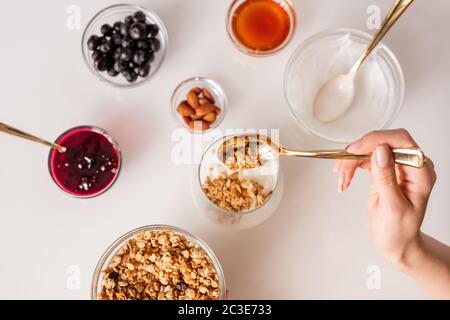 Hand of young woman with teaspoon putting muesli into glass with fresh sourcream while making yoghurt with jam, almond nuts, honey and berries Stock Photo