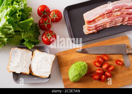 Still life composition consisting of two sandwiches, fresh tomatoes, lettuce, avocado and tray with bacon slices on kitchen table Stock Photo