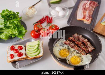 Two appetizing vegetarian sandwiches, frying pan with fried eggs and bacon, ripe fresh tomatoes, lettuce and hot chili peppers on table Stock Photo